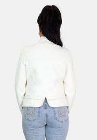 Chaqueta blanca de piel sintética con un diseño estructurado, espalda ventilada y sutiles detalles de costura, combinada con jeans adornados de un azul claro.