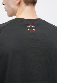 Μαύρο βαμβακερό μπλουζάκι με στρογγυλή λαιμόκοψη και μικρό κυκλικό κεντητό λογότυπο σε κόκκινο και λευκό στο πίσω μέρος. Λείο ύφασμα.