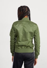 Veste bomber vert olive avec une texture lisse, des poignets et une ceinture côtelés. Comprend des épaules matelassées et un détail de poche latérale.