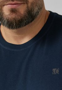 Homme barbu portant un t-shirt bleu marine à col rond avec un petit logo "s.O" sur la poitrine, visible du cou au haut du torse.