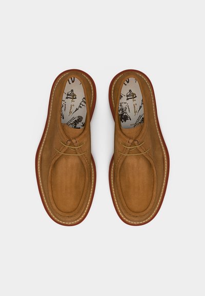 Ζευγάρι καφέ δερμάτινων αθλητικών παπουτσιών με κορδόνια, με αντίθετες κόκκινες σόλες και σχέδια στις εσωτερικές σόλες, εμφανιζόμενα από πάνω σε λευκό φόντο.