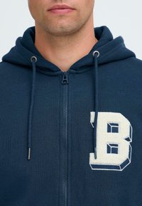 Muž nosí tmavomodrý mikinu na zips s kapucňou a veľkým bielym štruktúrovaným písmenom "B" na ľavej strane hrudníka.