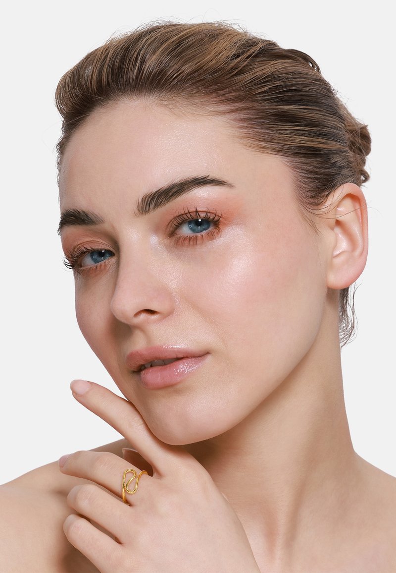 Goldener Ring mit einem gedrehten Design, der an einem Finger getragen wird. Das Model hat helle Haut, dunkles, nach hinten gebundenes Haar und zeigt dezentes Makeup in natürlichen Tönen.