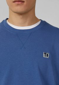 Uomo che indossa un felpa blu con colletto a girocollo sopra una maglietta bianca, mostrando l'area del petto e del collo con una piccola toppa con logo sul lato sinistro.