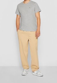 Uomo che indossa una maglietta a maniche corte grigia chiara, pantaloni beige con le mani in tasca e sneakers bianche e grigie, in piedi davanti a uno sfondo neutro.