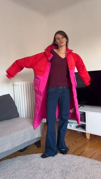 Casaco acolchoado vermelho e rosa com um design longo, apresentando dois grandes bolsos frontais e um capuz. Usado sobre um suéter roxo escuro e jeans.