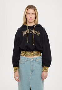 Hoodie nero cropped con "Just Cavalli" in strass, caratterizzato da una fodera a motivo floreale gialla e vita elastica. Abbinato a una gonna di denim chiaro.