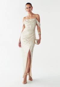 Zlata obleka z bleščečimi sequini, brezramenskimi rokavi in visokim razporkom na stegno; kombinirano z sandali s traki in majhno kovinsko torbico.