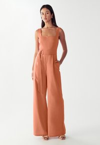 Žena oblečená v bezrukávovém, čtvercovém rustovém jumpsuitu se širokými nohavicemi, s uvázaným opaskem a sandály s otevřenou špičkou, stojící na bílém pozadí.