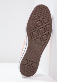 Semelle de chaussure Converse en caoutchouc brun avec motif de bande de roulement géométrique, soulignée en blanc, présentant une tige en toile orange clair et des œillets en métal.