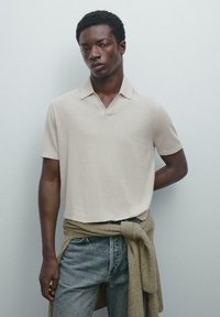 Giovane uomo che indossa una camicia beige a maniche corte con colletto, jeans blu e un maglione marrone annodato in vita su uno sfondo semplice.