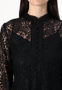 Μαύρη δαντελένια μπλούζα με περίπλοκα floral σχέδια, ψηλό λαιμό και κουμπιά κλεισίματος. Μακριά μανίκια με διαφάνεια και υφή.