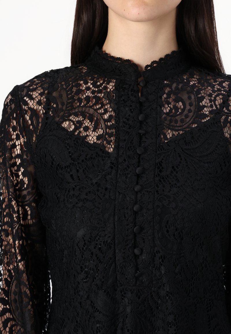 Schwarze Spitzenbluse mit kunstvollen floralen Mustern, hohem Kragen und Knopfverschlüssen. Lange Ärmel mit transparenter Optik und strukturiertem Finish.