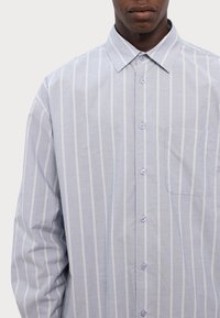 Camisa de botones azul claro con rayas verticales blancas, cuello apuntado, mangas largas y un bolsillo en el pecho izquierdo. La tela parece suave.