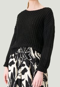 Pull noir en tricot avec une texture côtelée et de larges manches, associé à une jupe plissée noire et blanche à motifs floraux. Fini de surface mat.