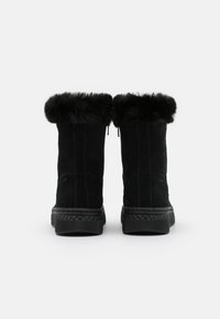 Une paire de bottes d'hiver en cuir suédé noir avec des poignets en fourrure, des semelles en caoutchouc épaisses et des coutures verticales, présentée de l'arrière sur un fond blanc.