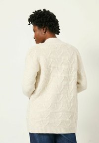 Cremfarbener, gestrickter Cardigan mit einem strukturierten Blütenblattmuster, langen Ärmeln und einem gerippten Saum. Über dunkelblauen Jeans getragen, von hinten gezeigt.