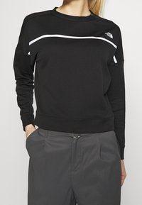 Persona che indossa una felpa nera con strisce bianche e il logo The North Face, abbinata a pantaloni grigi, mani in tasca, su uno sfondo neutro.