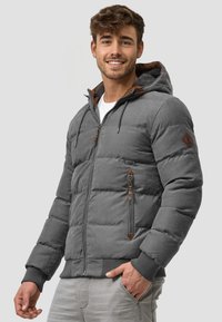 Grå puffjakke med hette, med teksturert overflate, brune detaljer, frontglidelås og ribbede mansjetter. Modellen har hendene i lommene.