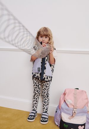 Barn med blondt hår iført bukser og vest med leopardprint leger med en metal slinky-legetøj ved siden af en pastel rygsæk på et gult tæppe.