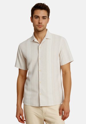 Junger Mann trägt ein kurzärmeliges, hellbeiges Hemd mit dezenten vertikalen Streifen, kombiniert mit cremefarbenen Hosen.