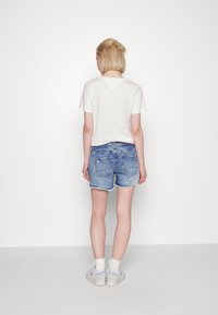 Henkilö, jolla on lyhyet vaaleat hiukset, seisoo valkoista seinää vasten, päällään valkoinen t-paita, siniset farkkushortsit, valkoiset sukat ja valkoiset tennarit.