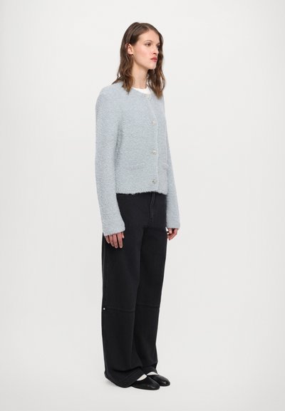 Lys blå teksturert cardigan med runde knapper, brukt over en hvit skjorte; kombinert med svarte vide bukser og flate sko.