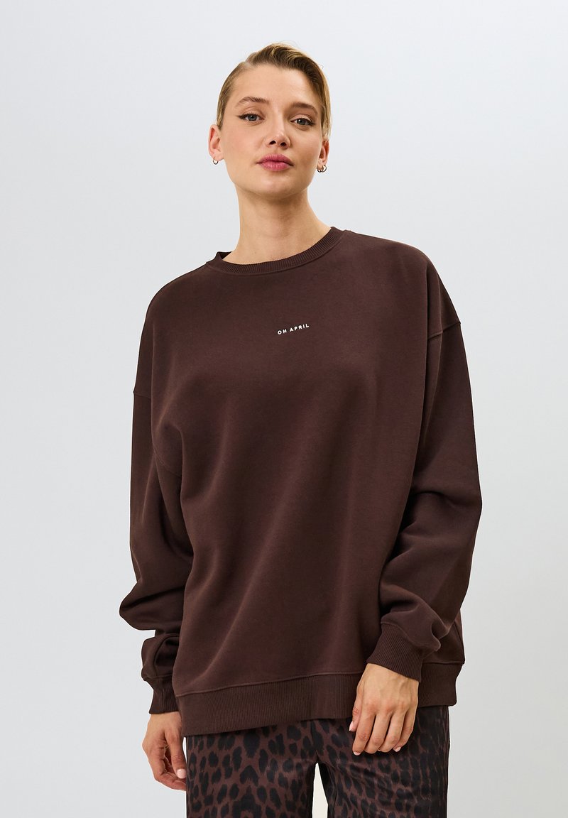 Hnedý bavlnený sveter s uvoľneným strihom, okrúhlym výstrihom a potlačou "OH APRIL" na hrudi, skombinovaný s nohavicami s leopardím potlačou.