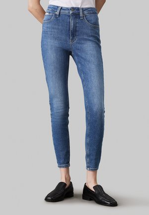 Magas derekú kék farmernadrág, testhez álló szabással, öt zsebbel és enyhe kopással, fekete slip-on cipőkkel párosítva.