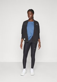 Zwarte sportjack over een blauwe top, gecombineerd met zwarte leggings en witte sneakers. Opvallende merklogo's op de kleding.
