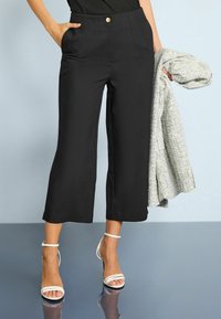 Pantalons noirs courts avec plis à l'avant et poches latérales. Portés avec des talons blancs à lanières et une veste grise texturée.