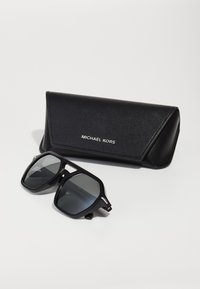 Ochelari de soare negri Michael Kors cu lentile întunecate, ramă rectangulară și accente argintii, alături de un toc asortat din piele sintetică neagră.