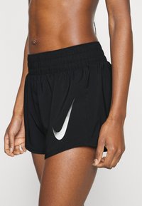 Pantalones cortos deportivos negros con una cinturilla ancha y elástica. Presenta un logo de Nike en plata reflectante en el costado y un diseño holgado.