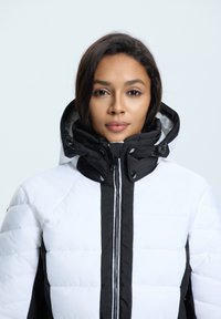 Witte pufferjas met een zwart voorpaneel en capuchon. Voorzien van een ritssluiting en drukknoopsluiting, met een doorgestikt patroon over het hele jasje.