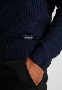 Navy blue textured knit sweater with a "YOURTURN" label, worn with one hand in the pocket of black pants.
