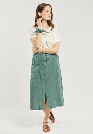 Femme debout, les bras croisés, portant un haut blanc à manches courtes, une jupe midi verte avec des poches avant, des sandales et un foulard vert au poignet.
