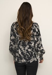 Blouse noire à motif floral avec manches longues bouffantes et coupe ample. Motif floral de couleur crème. Tissu lisse avec un léger éclat.