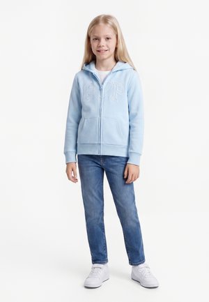 Ung pige står smilende iført lyseblå lynlåshættetrøje med hjerte-logo, blå jeans og hvide sneakers på hvid baggrund.