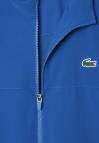 Giacca blu con zip e un piccolo logo di coccodrillo verde sul petto, con una cerniera parzialmente aperta che mostra il tessuto a rete all'interno.