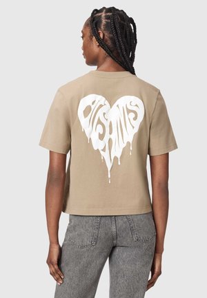 Person med fletninger, iført en beige t-shirt med et hvidt dryppende hjerte-design og grå jeans, vist bagfra.