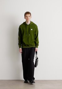 Hanorac verde cu buzunar frontal și șnur, asortat cu pantaloni negri și pantofi negri. Modelul ține o geantă mică neagră.