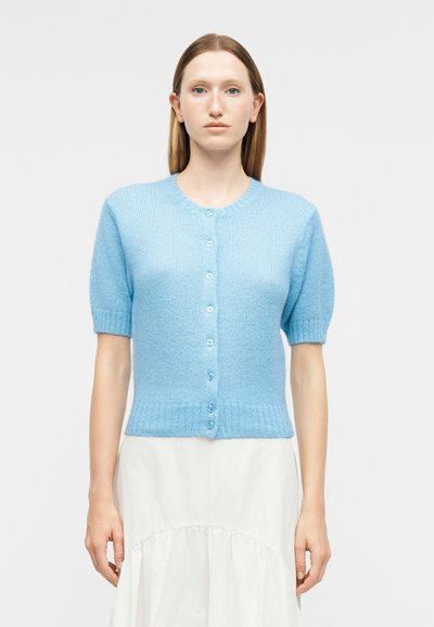 Femme portant un cardigan bleu clair à manches courtes boutonné et une jupe blanche, debout devant un fond uni clair.