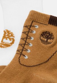 Une paire de chaussettes tricotées, l'une blanche et l'autre marron, toutes deux ornées d'un logo d'arbre et de détails de design de bottes à lacets.