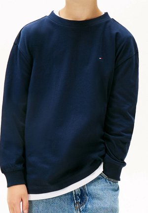 Person iført en mørkeblå langærmet skjorte over en hvid undertrøje, kombineret med lyseblå denim jeans, stående med den ene hånd i lommen.