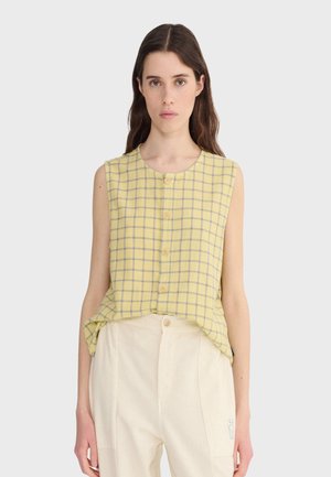 Žena oblečená v žltom károvanom topánke bez rukávov, zapnutej na gombíky a zastrčenej do vysokých svetlo-krémových nohavíc, stojaca pred jednoduchým pozadím.