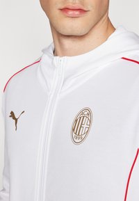 Hvid zip-up hoodie med røde detaljer, prydet med et guld Puma-logo og et guld AC Milan-emblem med året 1899. Blød stoftekstur.