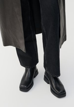 Schwarze Lederstiefeletten mit quadrater Spitze und klobiger Sohle, kombiniert mit dunkelblauen Jeans, die an den Knöcheln umgeschlagen sind. Glatte Textur und elegantes Design.