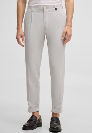 Hellgraue, maßgeschneiderte Hose mit Knopf- und Reißverschluss, umgeschlagenen Säumen, getragen mit schwarzen Leder-Loafern und einem weißen, in die Hose gesteckten Hemd.