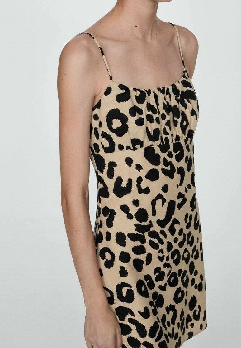Femme portant une robe beige sans manches avec fines bretelles et motif léopard noir irrégulier, torse et bras visibles sur fond uni.