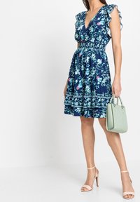 Vestido floral en azul marino con patrones verdes y rosas, con mangas con volantes, escote en forma de V y cintura fruncida. Combinado con un bolso de mano verde menta.